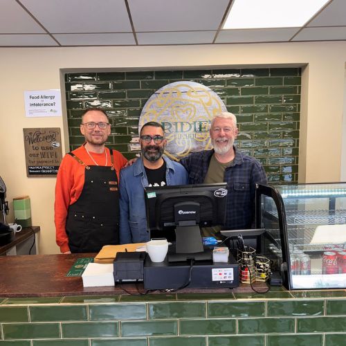 3 men behind a counter in a cafe smile at the camera